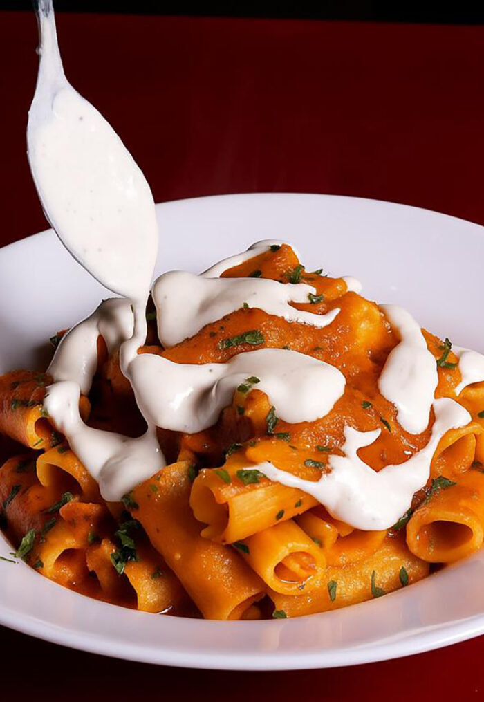 Rigatoni com Molho Cremoso
Neste prato, um garfo está adicionando um cremoso e espesso molho branco sobre um monte de rigatoni, uma massa em formato de tubo e de tamanho maior. A massa está envolvida em um molho à base de tomate com uma tonalidade alaranjada, que sugere a mistura com um laticínio, como creme de leite ou queijo.
O prato é finalizado com uma camada generosa do molho branco, que escorre sobre a massa. Há também uma guarnição de salsa picada espalhada sobre o molho de tomate, adicionando um toque de cor verde e um sabor fresco. Servido em um prato branco simples, o prato parece rico, reconfortante e convidativo.